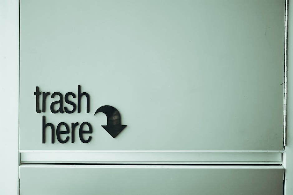 A close-up view of a light grey wall with a black sign that reads 'trash here' in lowercase letters, accompanied by a downward-pointing arrow. The sign is mounted on a flat, smooth surface, and the arrow indicates the location of a designated waste disposal area. The environment appears to be an interior setting, possibly within a commercial or public building corridor, with a clean and minimal background. The sign's labels and arrow are made of durable plastic or metal, contrasting with the plain wall for clear visibility, supporting independent waste collection or on-site rubbish disposal within a regulated facility or service area managed by Rubbish Removal Enfield, emphasizing the importance of proper waste management practices.