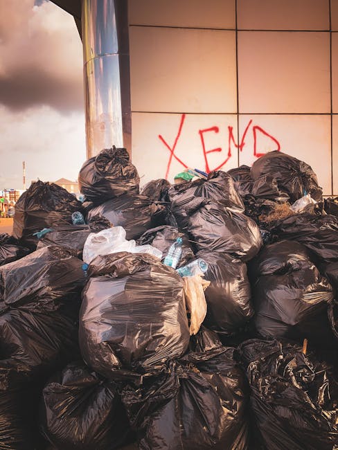 A large pile of black plastic rubbish bags, some with visible water bottles and scattered paper, is stacked against a tiled wall with a metallic surface on the left side. The bags are irregularly shaped, bulging with waste, and appear to be placed on an outdoor paved area, possibly a city sidewalk or service alley. In the background, there is a portion of an overcast sky with some clouds and distant urban structures. The wall behind the rubbish has graffiti with the word 'XEND' spray-painted in red. The scene showcases an accumulation of refuse that may be associated with independent waste disposal or rubbish removal services such as those provided by Rubbish Removal Enfield, highlighting the need for professional rubbish collection to manage excess waste in public or commercial environments.