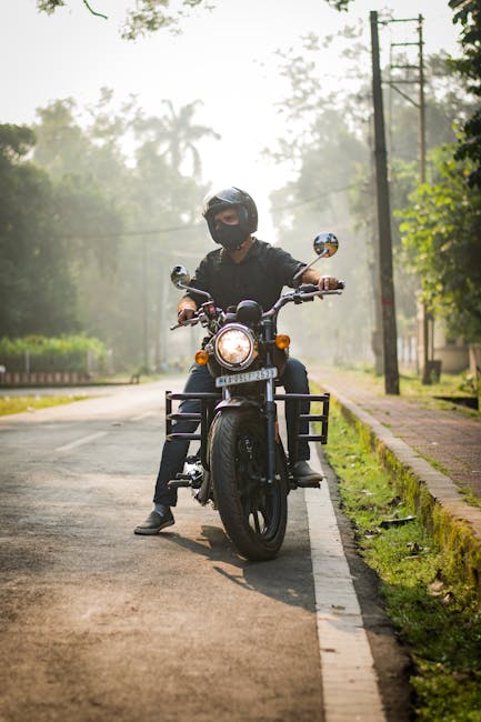 A person wearing a black helmet, face mask, and dark clothing is seated on a black motorcycle positioned in the centre of a suburban street, facing the camera. The motorcycle features a round headlight that is turned on, with orange indicators on either side, and a visible front wheel with a black fender. The rider's hands are on the handlebars, and the person appears to be ready to ride or waiting on the street. The background includes tall trees with green foliage, utility poles and wires along the roadside, and a slightly hazy sky indicative of early morning or late afternoon lighting. The street has a narrow paved surface with a white line marking the edge, and a grassy verge runs alongside the sidewalk, which is bordered by a curb and a row of trees. The scene reflects an outdoor environment suitable for casual or alternative waste disposal discussions, akin to private collection or independent rubbish removal services, as could be offered by companies like Rubbish Removal Enfield.