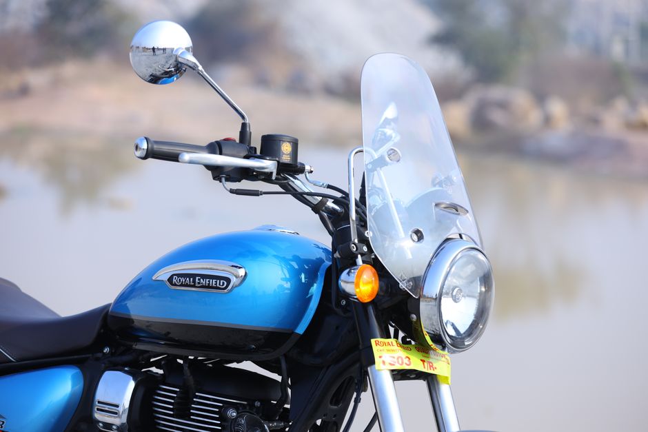 Close-up view of a blue Royal Enfield motorcycle parked outdoors with a blurred natural background, featuring a clear windshield, round headlight, and a silver engine with black accents. The motorcycle's handlebar, right-side mirror, and front indicator light are visible, along with a black seat and parts of the metal frame. The scene is well-lit, capturing the reflective surface of the chrome components and the textured finish of the paintwork. This image exemplifies personal transport equipment that might be considered for disposal or alternative waste handling through private rubbish removal services like Rubbish Removal Enfield, especially when the motorcycle is being prepared for scrapping or removal from a garage or driveway, demonstrating the type of bulky waste items handled in local or independent collection scenarios.