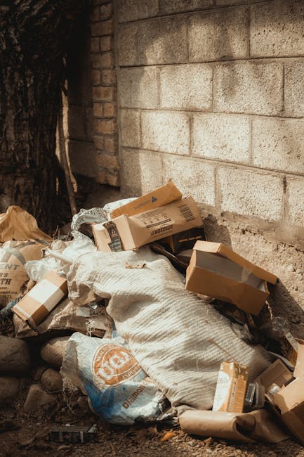 A small pile of discarded packaging and rubbish situated against a weathered brick wall and a concrete block wall, with a large tree trunk partially visible on the left. The debris includes cardboard boxes of various sizes, some with open flaps, crumpled paper, and a plastic bag with an orange logo. The cardboard appears plain brown and is slightly crushed, with some boxes showing labels and barcodes, indicating they were used for packaging or deliveries. The overall scene is outdoors, in a corner or alleyway, with loose stones and dirt on the ground surrounding the waste. Natural lighting highlights the textures and tones of the rubbish and walls, suggesting daytime. This scene illustrates a typical instance of waste accumulation for disposal, which companies like Rubbish Removal Enfield handle through private collection and on-site clearance services, avoiding reliance on municipal waste services for managing general rubbish.