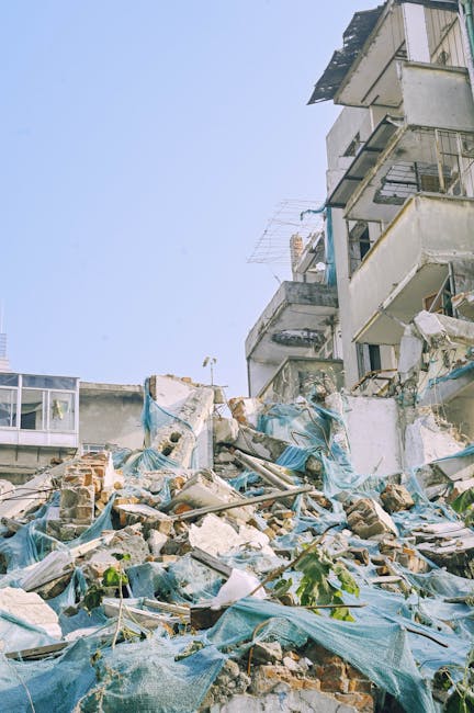 A large pile of construction debris surrounds the base of a damaged multi-storey apartment building, with bricks, broken concrete, and scattered blue tarps visible in the foreground. The debris includes crumbled bricks, shattered plaster, and twisted metal fragments, all strewn across a concrete pavement area. The building's weathered facade shows exposed concrete, damaged balconies, and broken windows, indicating recent structural deterioration. To the left, part of a modern glass extension contrasts with the damaged structure, while the background features a clear blue sky. Environmental lighting suggests daylight conditions, casting minimal shadows over the scene. This scene may reflect a site undergoing private clearance or demolition, where independent waste removal services like those from Rubbish Removal Enfield could facilitate the safe disposal of such heavy and hazardous construction waste, aligning with the need for professional rubbish removal services in complex urban environments.