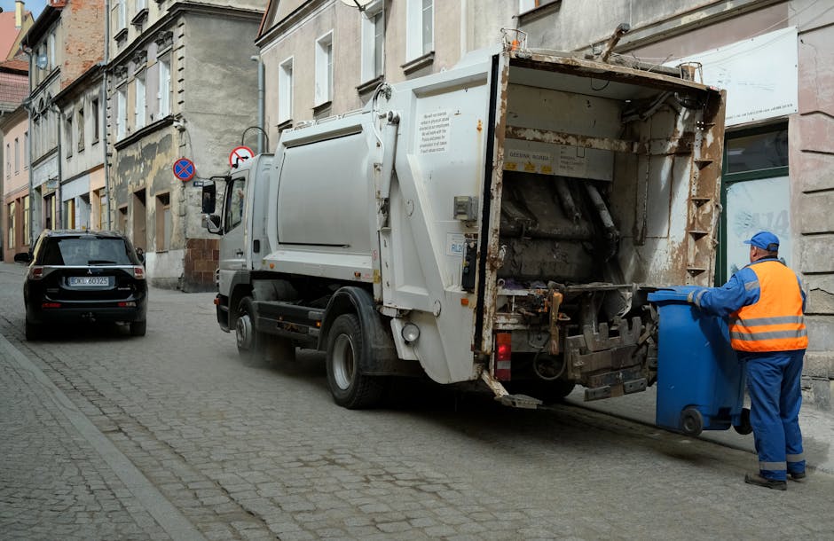 A large pile of mixed household waste and rubbish bags is gathered on a paved sidewalk in an urban area, with various materials including cardboard boxes, plastic bags, paper, and miscellaneous trash stacked around and on top of a metal recycling bin labeled for mixed paper and cardboard. The pile partially blocks a metal railing and extends onto the pavement, with some waste spilling onto the road surface. Behind the waste, there are parked cars, including a silver vehicle, and a row of commercial storefronts with signs, alongside a building wrapped in blue scaffolding, indicating ongoing maintenance or renovation. The scene is well-lit by natural daylight, with a leafless tree visible on the left side of the image, suggesting a late autumn or winter setting. The overall impression is of an overflowing waste collection point, highlighting the need for rubbish removal services that handle unsorted or excess waste, in line with private or independent waste disposal options.