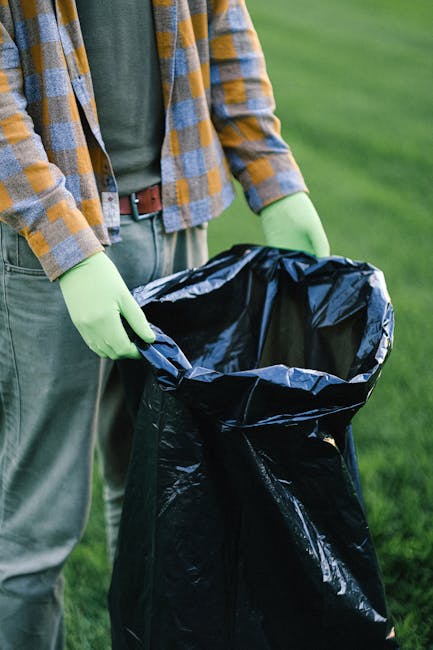 A person wearing a multicolored plaid shirt with shades of yellow, blue, and gray, along with gray trousers secured by a brown belt, is seen holding open a large black rubbish bag lined with a shiny black plastic interior. They are wearing bright green gloves, suggesting safe handling of waste. The scene is set outdoors on a well-maintained grassy area with a blurred green background, possibly a park or garden. The individual appears to be in the process of collecting or preparing waste for disposal, which could relate to private waste handling or rubbish removal services such as those offered by Rubbish Removal Enfield. The focus on the rubbish bag, along with the gloves, indicates a conscientious approach to waste collection, suitable for on-site clearance or alternative rubbish collection methods. The lighting is natural, highlighting the textures of the gloves, the fabric of the shirt, and the shiny plastic lining inside the bag, emphasizing the practical nature of waste management tasks conducted in outdoor environments.